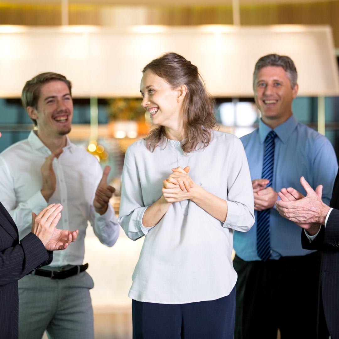 Closeup of four smiling people applauding and congratulating business woman and standing around her with blurred view in background.